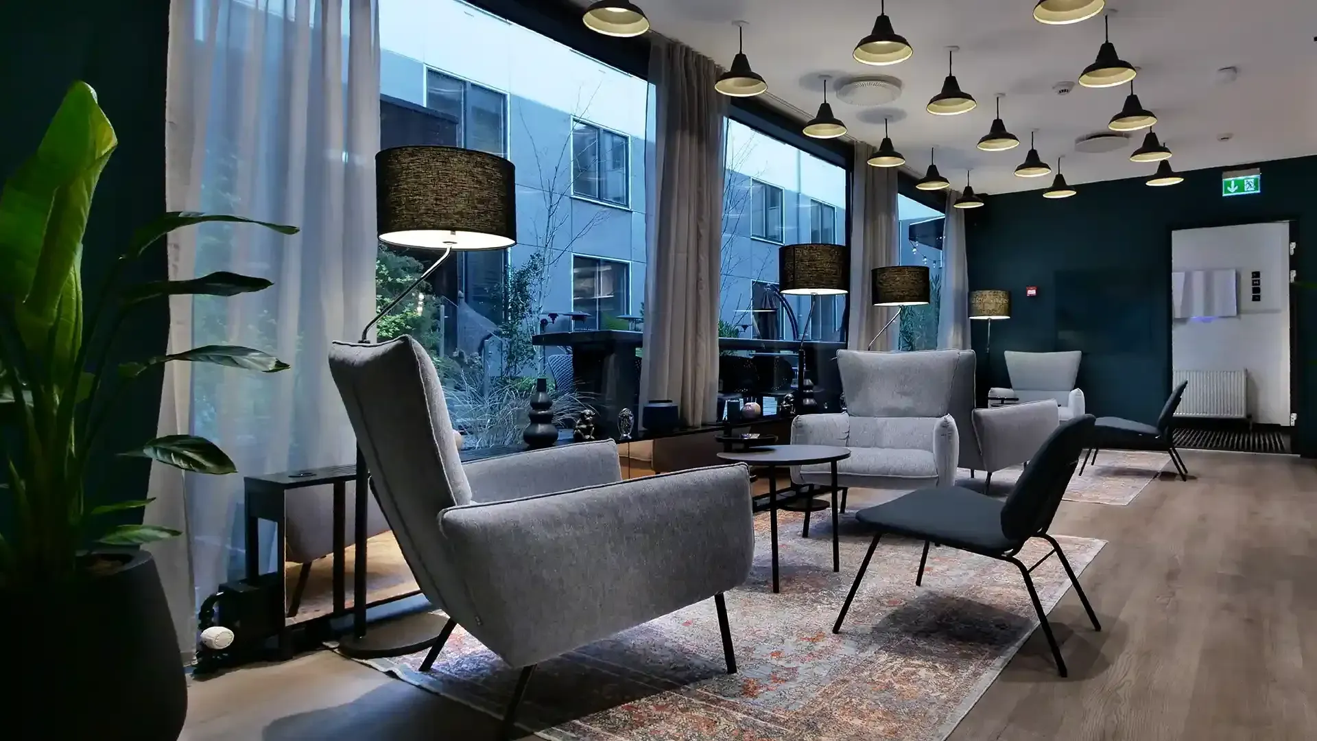 Light-filled lounge with grey chairs, large windows and graphic ceiling lamps – modern, calm and functional hotel interior.